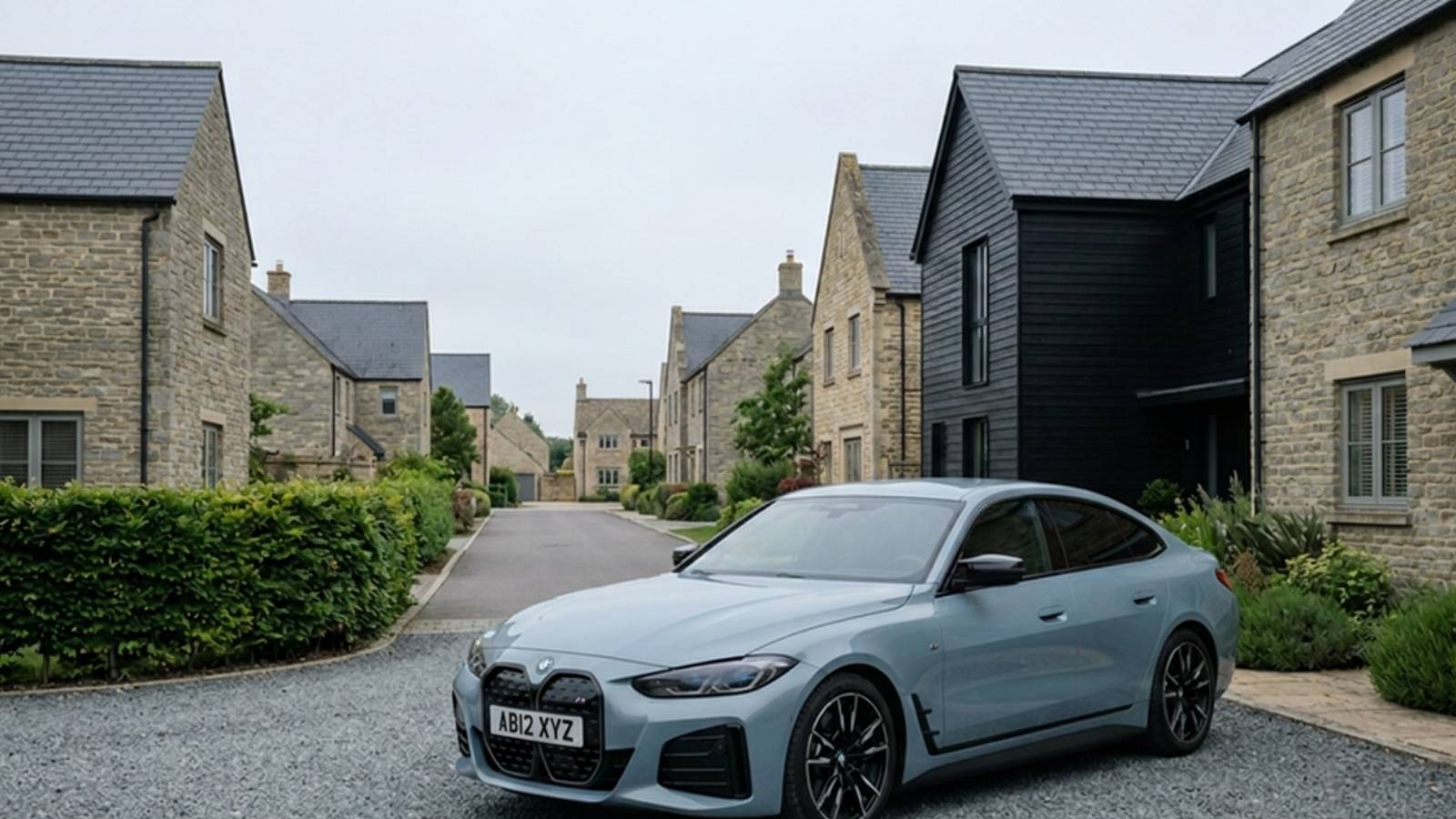 A sky-grey BMW i4 Gran Coupé on a gravel driveway in a Cotswold-stone housing development, seen front three-quarter, showing its five-door liftback silhouette.