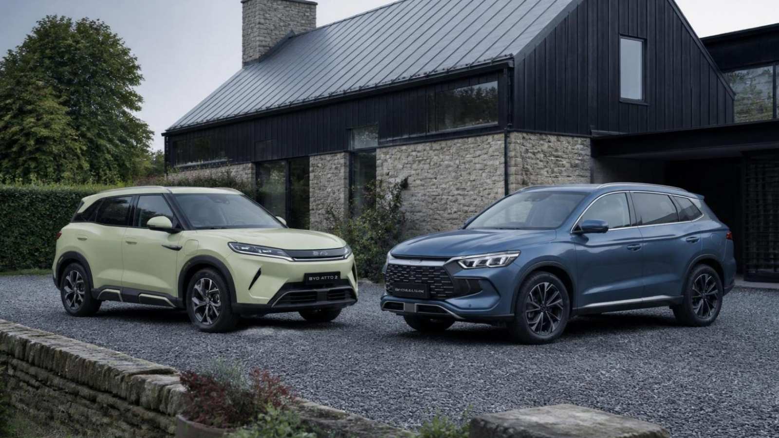 Two BYD electric SUVs, a pale lime-green Atto 2 and a blue Sealion, parked on gravel in front of a barn with black timber cladding and stone walls, under an overcast sky.