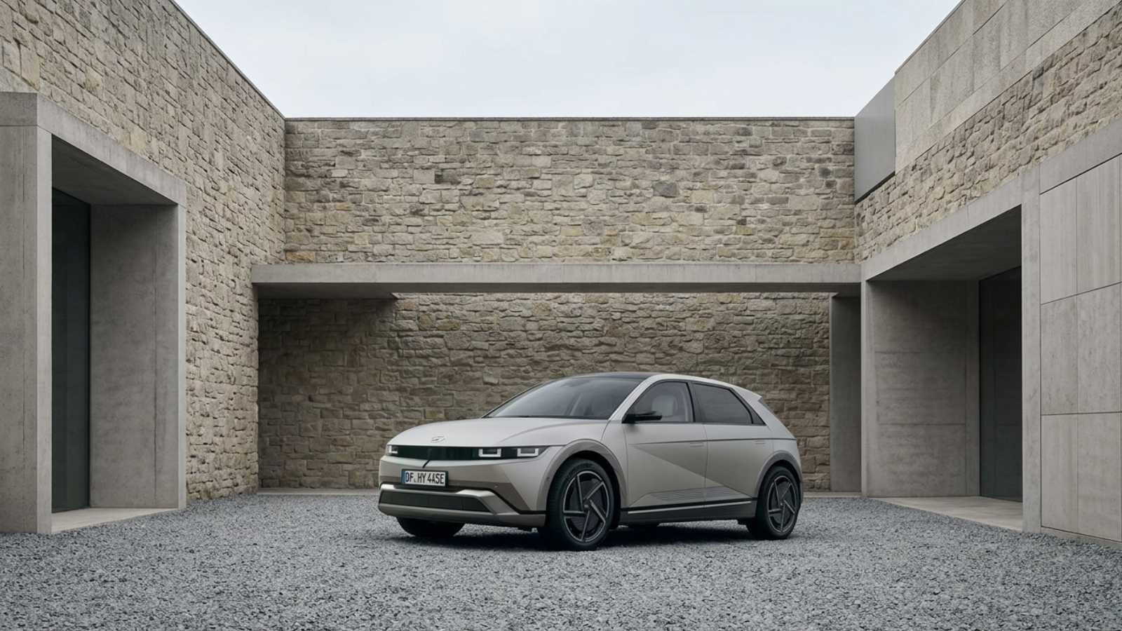 A sand-grey Hyundai Ioniq 5 seen front three-quarter on a gravel courtyard before stone and concrete walls, showing its upright crossover stance and pixel lighting.
