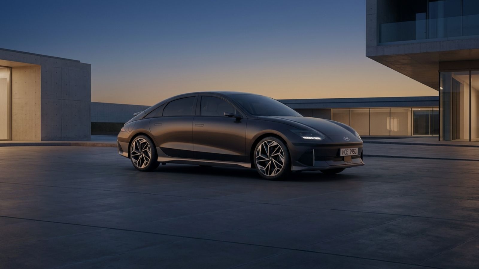 A dark metallic Hyundai Ioniq 6 saloon seen from the front three-quarter angle on a concrete plaza at dusk, showing its low streamliner roofline, ducktail and slim light bar.