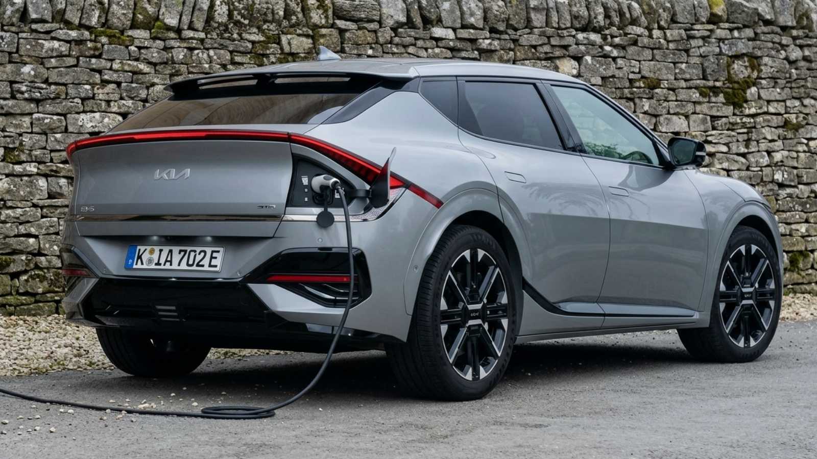 A silver Kia EV6 electric crossover seen from the rear three-quarter beside a drystone wall, plugged in and charging, showing its full-width rear light bar and fastback roofline.