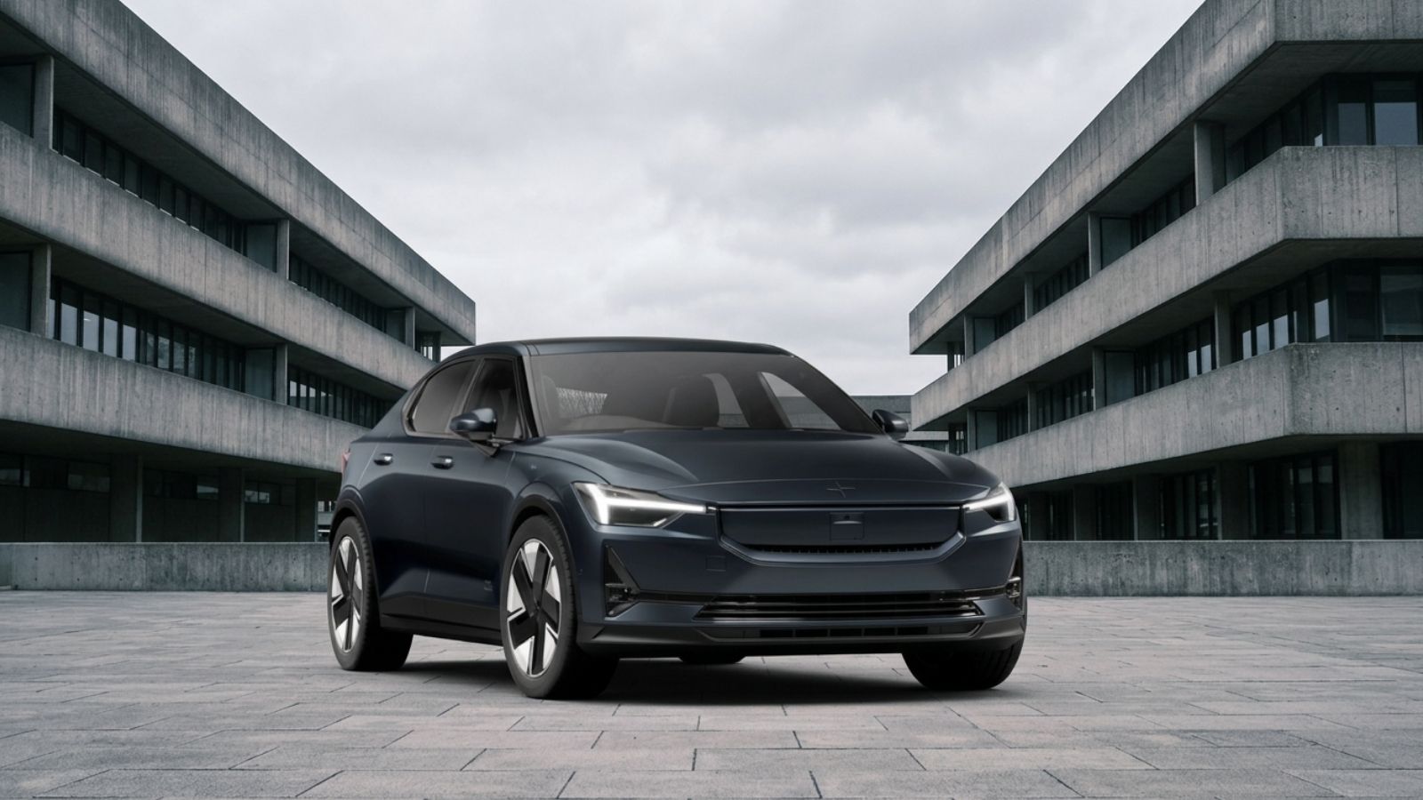 A dark grey Polestar 2 electric fastback in front three-quarter view on a paved plaza between grey concrete buildings, showing its blanked SmartZone front and LED headlights.