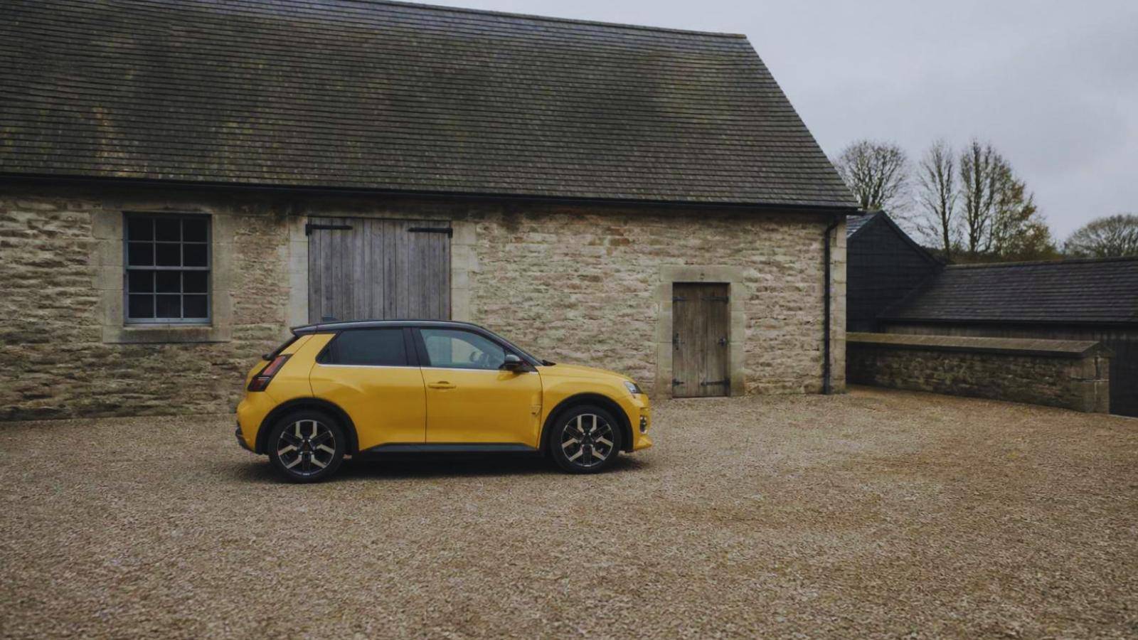 A Renault 5 E-Tech parked on a UK residential street in overcast daylight, a charging cable run to a wall-mounted home charger on the brick frontage.