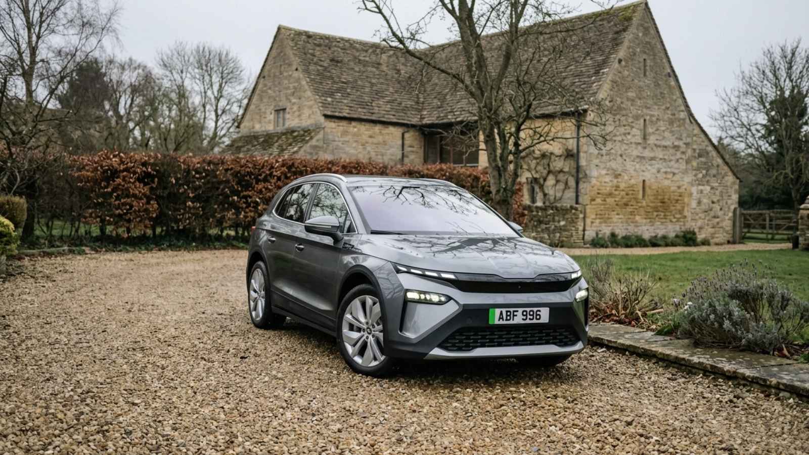 A grey Skoda Enyaq electric SUV seen front three-quarter on a gravel driveway before a Cotswold-stone barn, showing the Modern Solid Tech-Deck front and split LED lights.