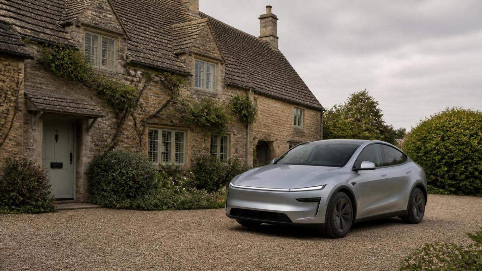 A silver 2026 Juniper Tesla Model Y with its full-width front light bar, parked on a gravel driveway in front of a Cotswold-stone cottage.