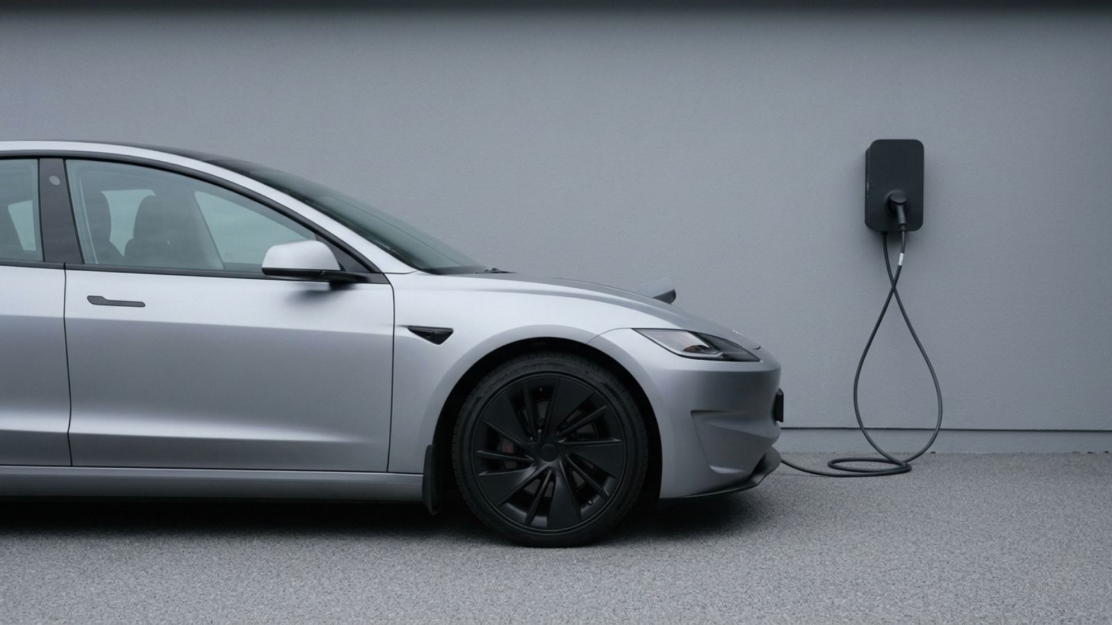 A silver Tesla with black alloy wheels charging from a wall-mounted home charger on a grey forecourt