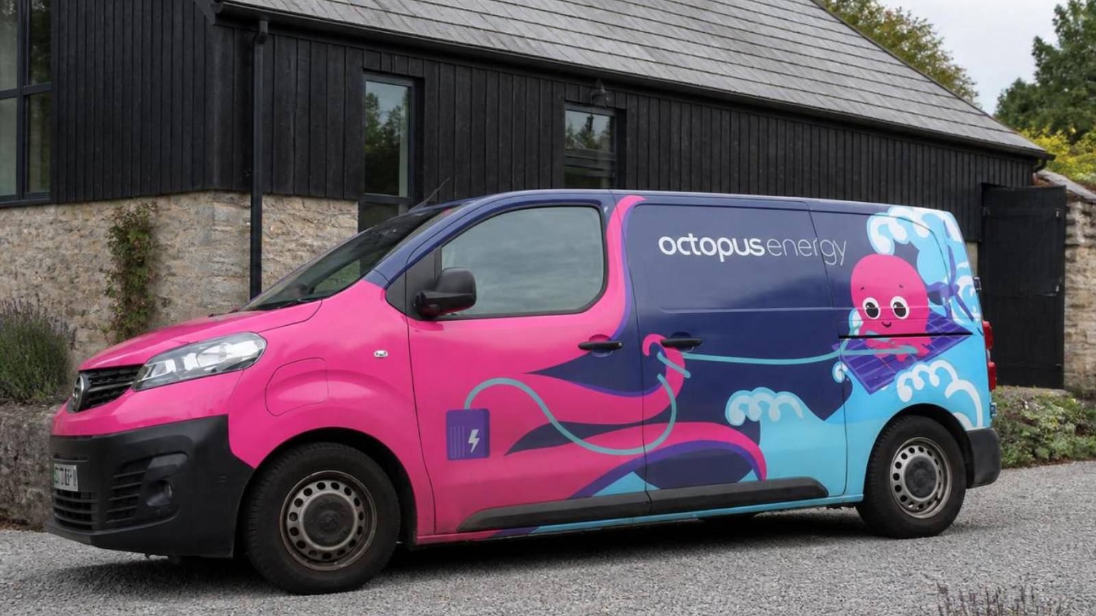 An Octopus Energy-branded pink and blue electric van parked on a gravel forecourt in front of a modern black-timber and stone UK building on an overcast day.